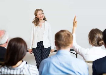 Smiling young businesswoman leaning on table and looking at woman from audience raising hand. Rear view of audience of five people.