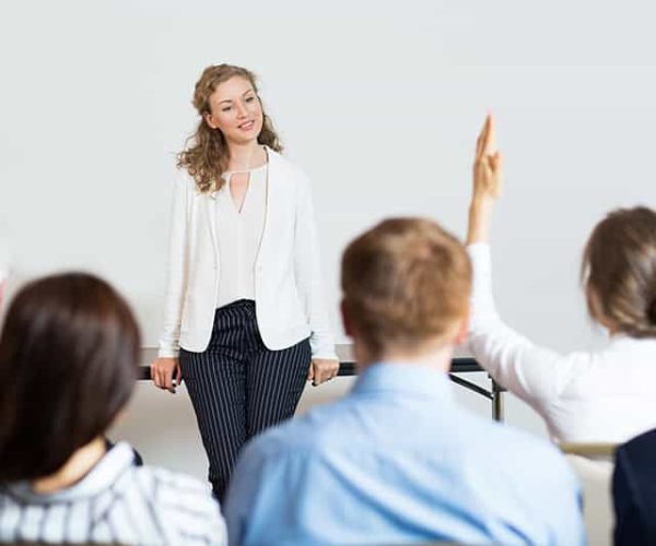 Smiling young businesswoman leaning on table and looking at woman from audience raising hand. Rear view of audience of five people.